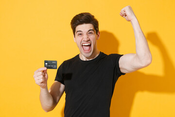 Screaming young man guy in casual black t-shirt posing isolated on yellow background studio portrait. People emotions lifestyle concept. Mock up copy space. Hold credit bank card doing winner gesture.