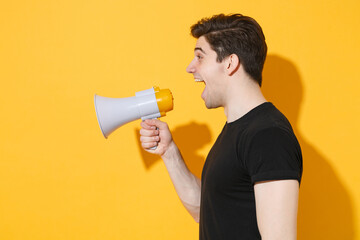 Side veiw of shocked young man guy in casual black t-shirt posing isolated on yellow background studio portrait. People sincere emotions lifestyle concept. Mock up copy space. Screaming in megaphone.