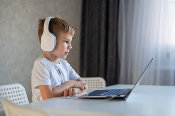 Cute diligent boy is typing on the keyboard and is carefully looking at the laptop screen. Caucasian schoolboy sits at a table, listens to music on headphones and does homework. Distance education.