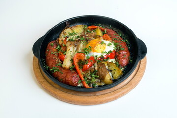 Dish on a black pan, which stands on a wooden round stand. Baked potatoes with sausages and red pepper. Sprinkled with green parsley.