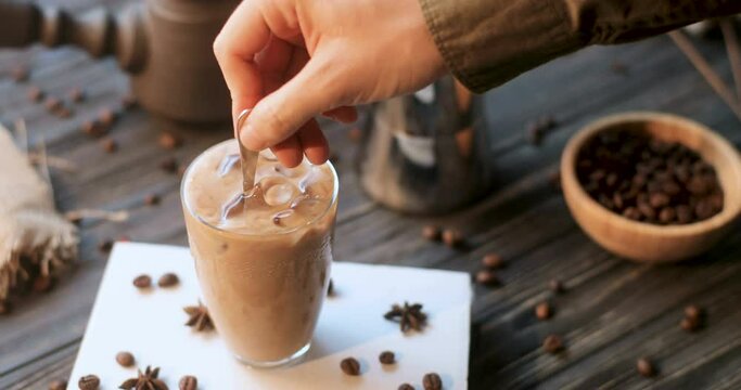 Close up of male Caucasian hand putting spoon in glass of frappe with ice cubes and mixing. Man barrista making cold coffee. Table in cafe with coffee seeds.