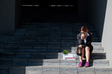 Depressed woman dressed in medical mask is fired and is sitting on the stairs with a box of personal belongings. Female office worker in suit and sneakers outdoors. Unemployment in the economic crisis