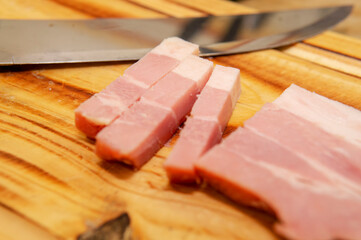 A  sliced bacon next to a shiny metal knife placed on top of a wooden cutting board. Taken indoor under a soft white light
