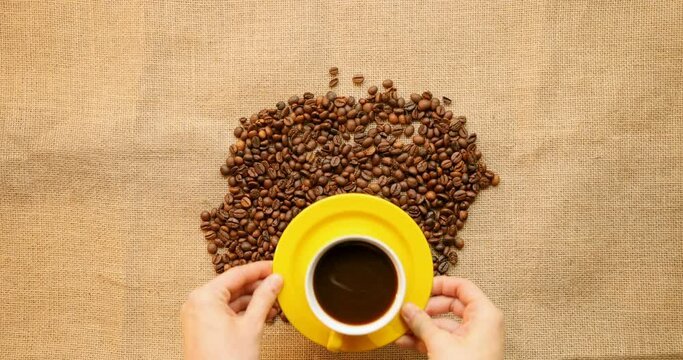 Flat lay on Caucasian male hand putting yellow cup of hot coffee drink on roasted beans and linen cloth. Top view on cafe advertisement composition on table.