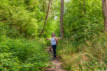 Mature Mexican woman with a surprised expression with her dog on a dirt road surrounded by trees and green foliage vegetation in the middle of the forest, sunny day in South Limburg, the Netherlands