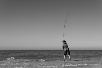 Black and white photo of a fisherman wearing a hat throwing his long curved fishing rod into the...