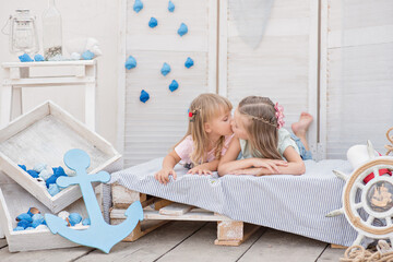 The best sisters in the world! Two little blonde girls play with shells, stars, an anchor, lanterns and marine decor, lie on the bed, laugh, hug, kiss, listen to the sounds of the sea, share secrets