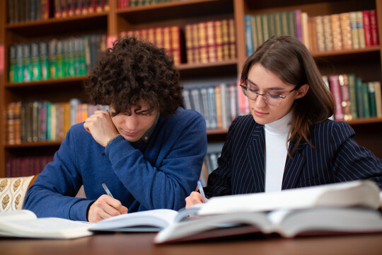 University students, a curly guy in a blue sweater and a brunette girl in glasses in a black jacket doing their homework in the library. Students learn hard.
