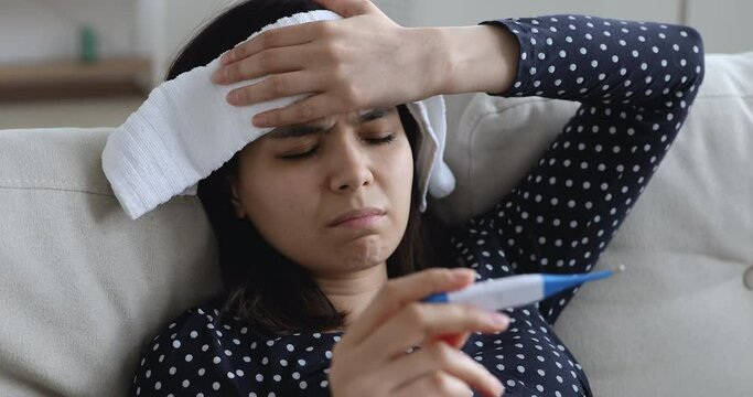 Close up Asian woman hold electronic thermometer check body temperature feels sick put piece of wet towel on forehead to reducing high fever. Covid-19 corona virus or seasonal grippe symptoms concept