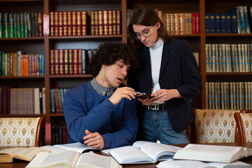 A curly-haired student in a grey-collared sweater sits at a table and explains a brunette girl standing next to her in glasses in a black jacket, against the backdrop of bookshelves in the library