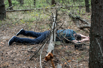 A body in the woods. A dead man in a blue t-shirt and trousers is pinned down by a fallen tree. Concept of accidents in the forest.