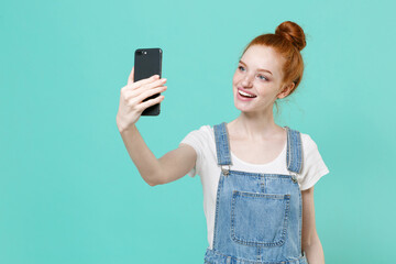 Smiling young readhead girl in casual denim clothes posing isolated on blue turquoise wall background studio portrait. People lifestyle concept. Mock up copy space. Doing selfie shot on mobile phone.