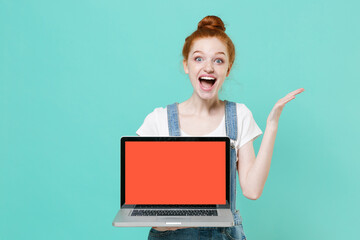 Naklejka premium Excited young readhead girl in casual denim clothes isolated on blue turquoise background. People lifestyle concept. Mock up copy space Hold laptop pc computer with blank empty screen spreading hands.