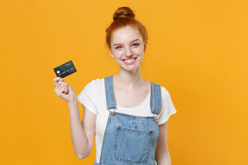 Smiling young readhead girl in casual denim clothes white t-shirt isolated on yellow background studio portrait. People sincere emotions lifestyle concept. Mock up copy space. Hold credit bank card.