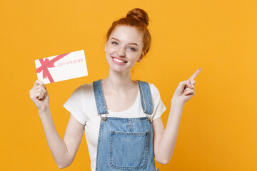 Smiling young readhead girl in casual denim clothes white t-shirt isolated on yellow background. People lifestyle concept. Mock up copy space. Hold gift certificate, pointing index finger aside up.