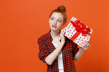 Pensive young readhead girl in checkered shirt posing isolated on orange background. St. Valentine's Day International Women's Day birthday holiday concept. Hold red present box with gift ribbon bow.