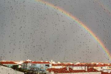 Double rainbow over buildings seen through window with raindrops