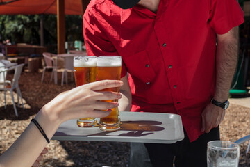 Waiter with protective mask on face serving beer to client at outdoor bar