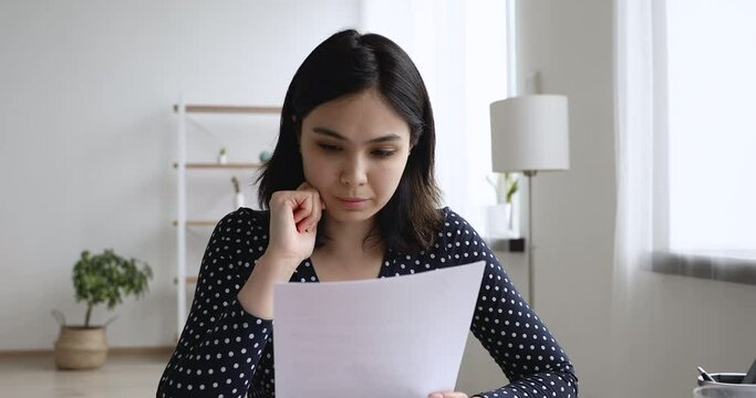 Asian young student girl sit at desk at home prepare essay read report document ponders over amendments feels doubtful insecure about paragraph looks out the window thinking study work process concept