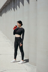 A taut Asian woman in a black tight-fitting tracksuit stands near a concrete wall and drinks water from a red drinking bottle outdoors in a park