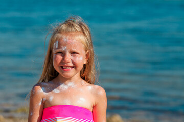 A beautiful little girl smiles in a swimsuit standing by the sea with sunscreen smeared on her face.