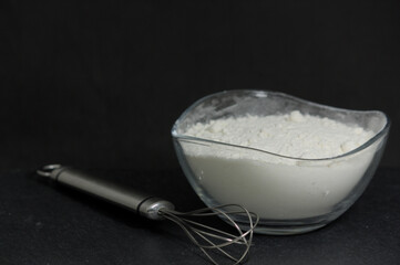 White flour in a bowl next to a silver whisk on dark background