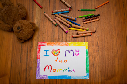 Child painting on a wooden table reading; "i love my two moms" in a rainbow frame, colorful crayons and a teddy bear around it