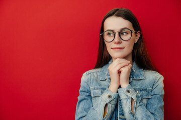 Portrait of pretty calm young woman in eyewear keeps hands together near face, looks optimistic aside, dressed in denim jacket, hears pleasant story from friend, isolated over red studio background.