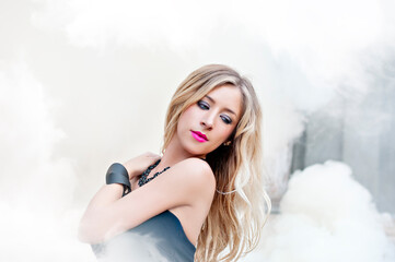 Beautiful woman model with long blond hair in a black evening dress with open neckline decoration on the neck, the bracelet on arm and pink lips standing in the smoke from the bombs on the background