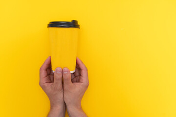 Two caucasian male hands hanging yellow paper cup with black lid with coffee take away on yellow background, copy space.
