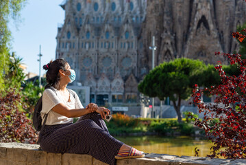 young and pretty tourist woman with face mask posing in front of sagrada familia in barcelona,...