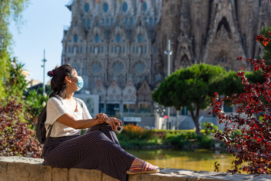 Young And Pretty Tourist Woman With Face Mask Posing In Front Of Sagrada Familia In Barcelona, Symbol Of Tourism To Spain During The Covid Disease And Corona Virus Pandemic