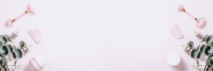 Facial roller, gua sha and moisturizing cream with green leaves. Natural cosmetics and facial tools on pink, web banner. Rose quartz roller, gua sha flat lay. Skin care routines, top view, copy space