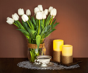 Bouquet of white tulips in a vase with burning candles and cup of tea on the table. Moody atmosphere.