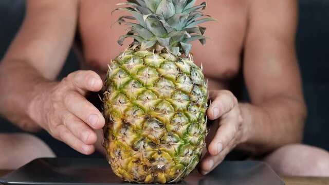 tanned old man twists from all sides in his hands a ripe yellow pineapple, the concept of vegetarian food, relaxation in exotic countries