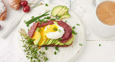 toast with poached egg and avocado on a round board, next to croissants and ripe red cherries
