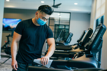 young asian male wearing mask, cleaning treadmill with sanitizing wipe COVID19