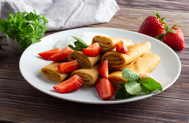 Pancakes with fresh strawberry, mint and cream on white plate. Brown wooden background, close up. Traditional breakfast.