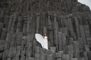 Destination Iceland wedding. The bride in a white silk dress with a bouquet in her hands, stands on black sand, on the beach of Vik, under a rocky mountain of basalt stones.