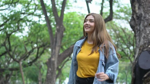 HD Slow motion low angle view of smiling young beautiful Asian woman with long hair looking up at the tree at the park in summer. Happy pretty teenage girl enjoy and having fun in beautiful nature.