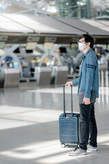 Asian man tourist wearing face mask carrying suitcase luggage waiting for check-in in airport terminal. Coronavirus (COVID-19) pandemic prevention when travel. Health awareness and social distancing