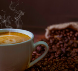 Hot fresh coffee with smoke and milk foam in a white ceramic cup with coffee beans roasted in sack bag on table background.