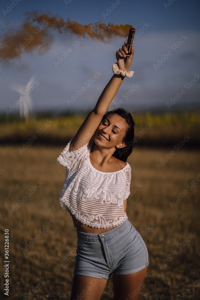 Poster vertical shot of a caucasian female posing with smoke bomb on background of fields and windmills