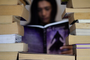 Woman with rows of books in front of her, with her face appearing in the middle of the books. A square made of books with a woman in the background trying to get a book out of the amount of books.