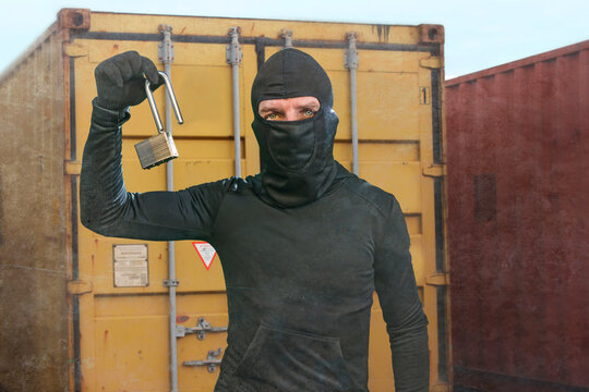 thief at work - criminal man in black covered with balaclava mask holding unlocked padlock at shipping area break in storage containers in robbery and crime concept