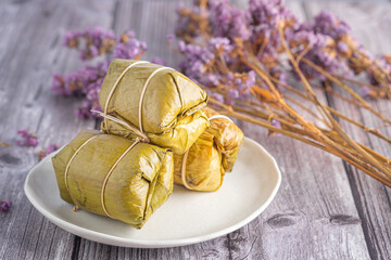Steamed sticky rice with banana wrapped banana leaf on plate with purple flowers blurred background.