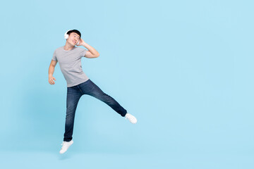 Fototapeta na wymiar Smiling energetic young Asian man wearing headphones listening to music jumping and looking at copy space on light blue isolated background