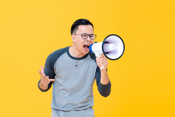 Angry young Asian man in gray top and eyeglasses shouting into megaphone isolated on yellow...