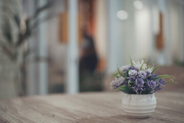 Close up of small purple flower on a white pot with blurry cafe ambience as background.