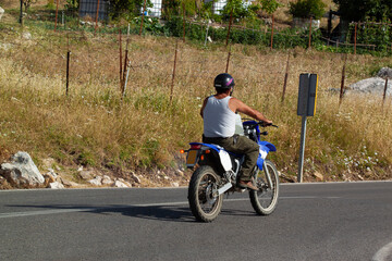 A spanish villager is riding a blue motorbike among farmlands, while carrying a barrel in front of him. He wears white underwear vest and a helmet. His arms are tanned due to direct sunlight.